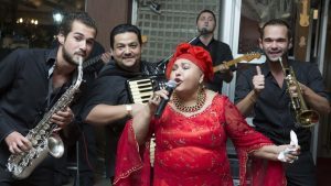 Joyful musical ensemble performing live with passion, featuring a vocalist in a vibrant red outfit, saxophonist, accordionist, and trumpeter.