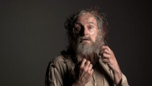 An elderly man with a long beard and tangled hair, embellished with twigs, looks on with an expression of surprise or concern.