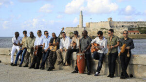A group of musicians with various instruments, including guitars and percussion, gathered along a coastal wall with an old lighthouse and fortification in the background, evoking a sense of cultural heritage and camaraderie.