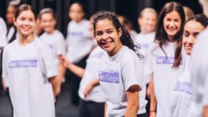 A group of smiling young performers in matching t-shirts gather on stage, radiating excitement and camaraderie during a dress rehearsal or event.