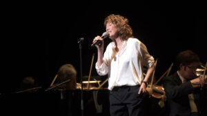 A vocalist performing on stage with a microphone, accompanied by a pianist and other musicians in an orchestral setting.