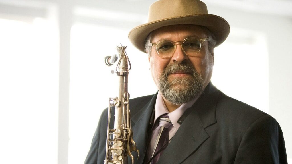 A distinguished gentleman with a beard and glasses, wearing a fedora hat, a tie, and a suit, holding a saxophone and smiling confidently.