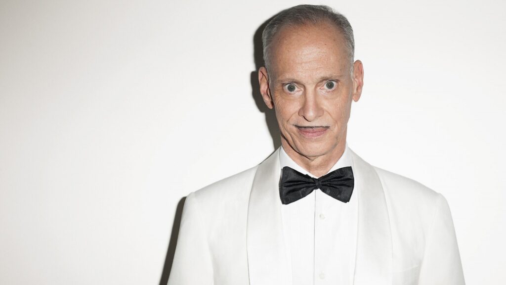 A man in a white tuxedo and black bow tie poses against a plain background, giving a distinctive look with a hint of charisma.