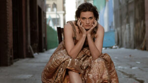 Madeleine Peyroux in a contemplative pose, dressed in an elegant gown, sitting on a deserted urban street.