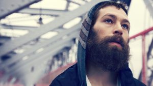 Matisyahu with a contemplative expression gazes into the distance, wearing a hooded jacket in an industrial setting.