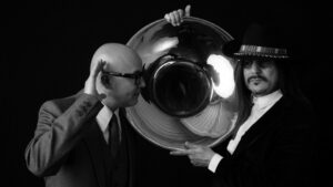 Two stylish men in monochrome attire facing each other, one gesturing toward a large, reflective tuba with a sense of intrigue, adding a touch of surrealism to a classic portrait.