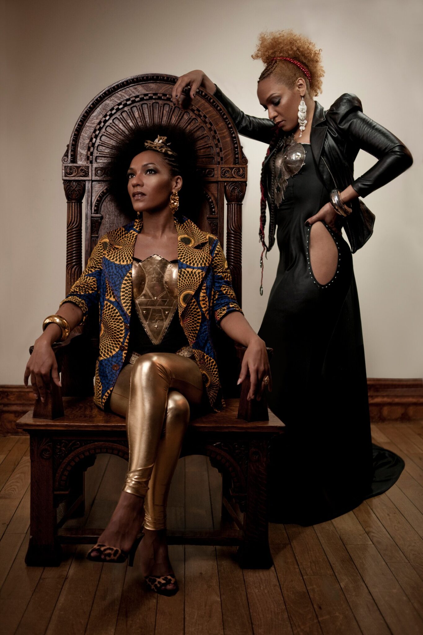 Two women exuding confidence and regality, with one sitting on a throne-like chair and the other standing beside her, both dressed in outfits with african-inspired elements and bold accessories.