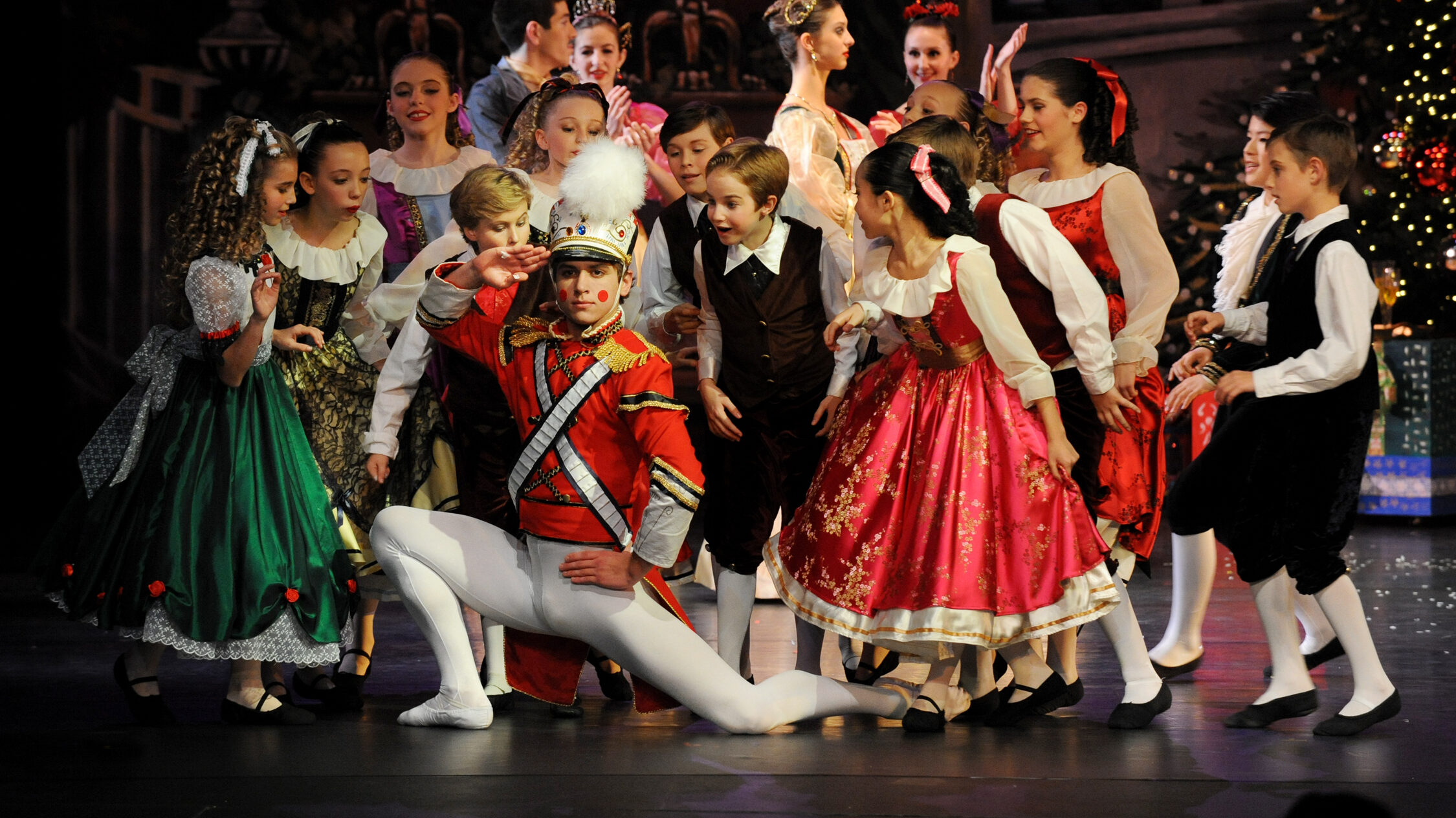 A group of children dancers in colorful, festive costumes performing a scene from "the nutcracker" ballet, with a young performer in a soldier costume sitting center stage surrounded by his peers.