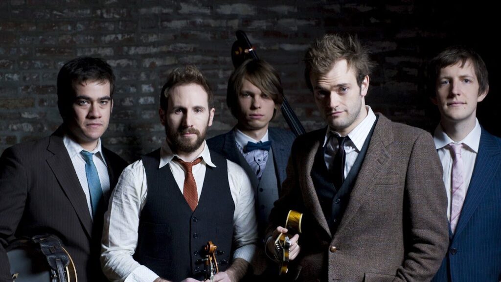 Five musicians posing confidently with their instruments against a rustic brick backdrop, exuding a cool, indie-band vibe.
