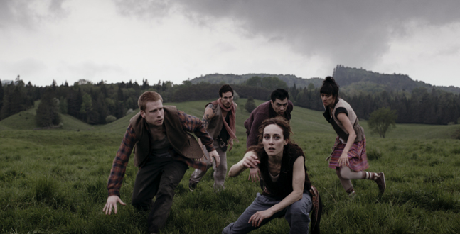 A tense moment in a grassy field, where a group of people appear to be intensely focused or alarmed, with some crouching and others poised to run, against a backdrop of overcast skies and rolling hills.