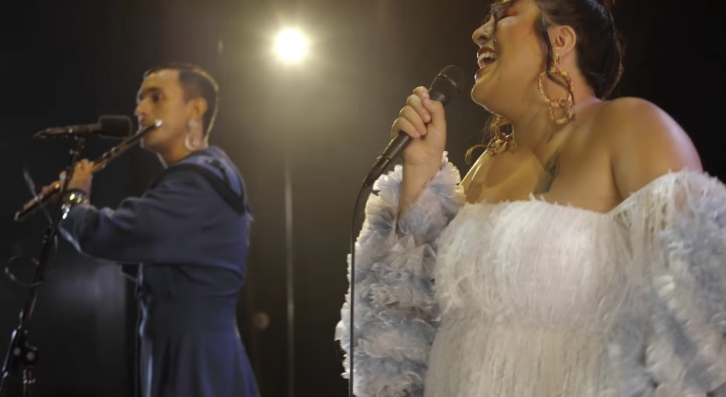 A duo immersed in a musical performance, with San Cha taking center stage as she sings passionately into the microphone, while a male musician in the background contributes with his instrument under the warm glow of a stage