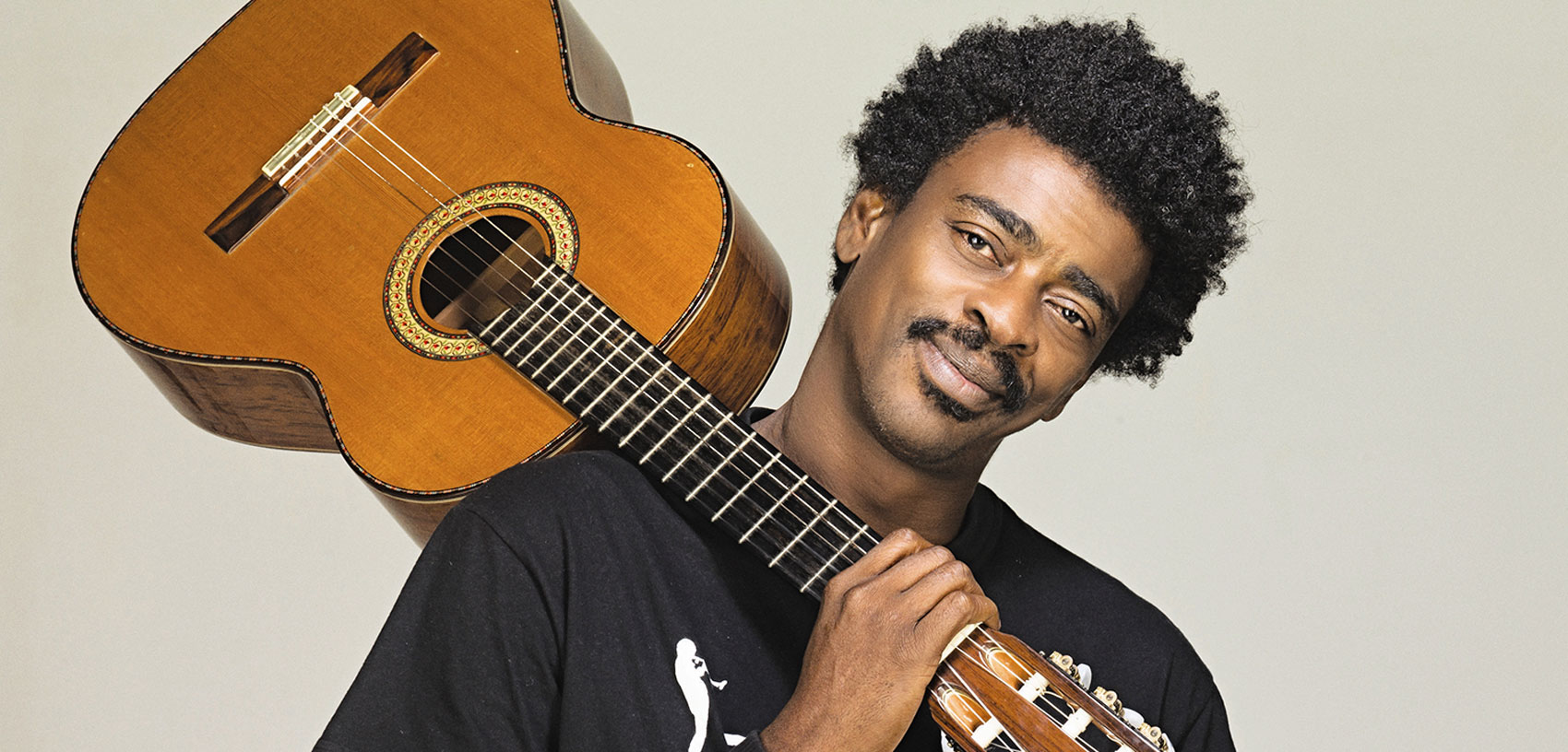 Man with an afro smiling as he balances an acoustic guitar on his shoulder.