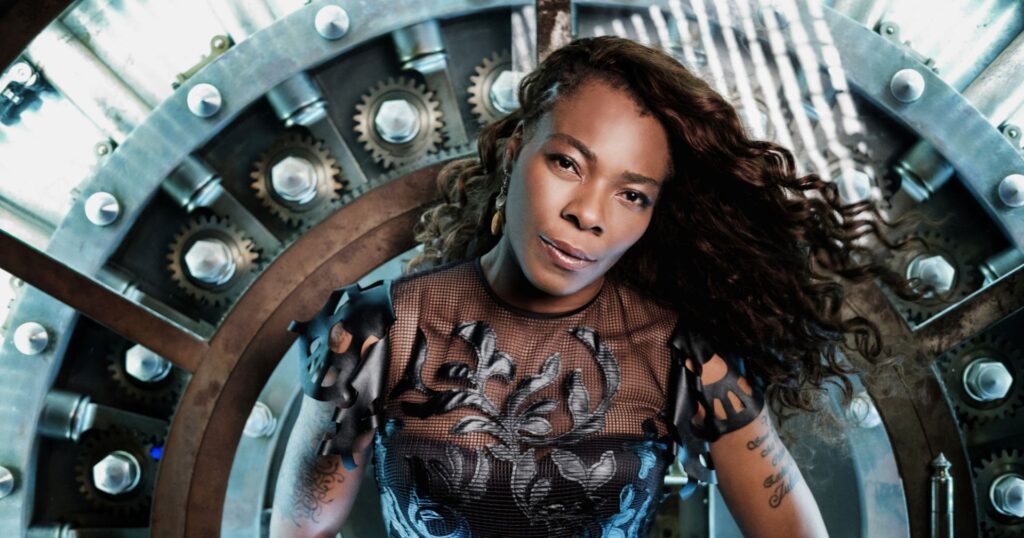 A confident woman in a stylish outfit poses in front of a mechanical, gear-filled backdrop, exuding a futuristic steampunk vibe.