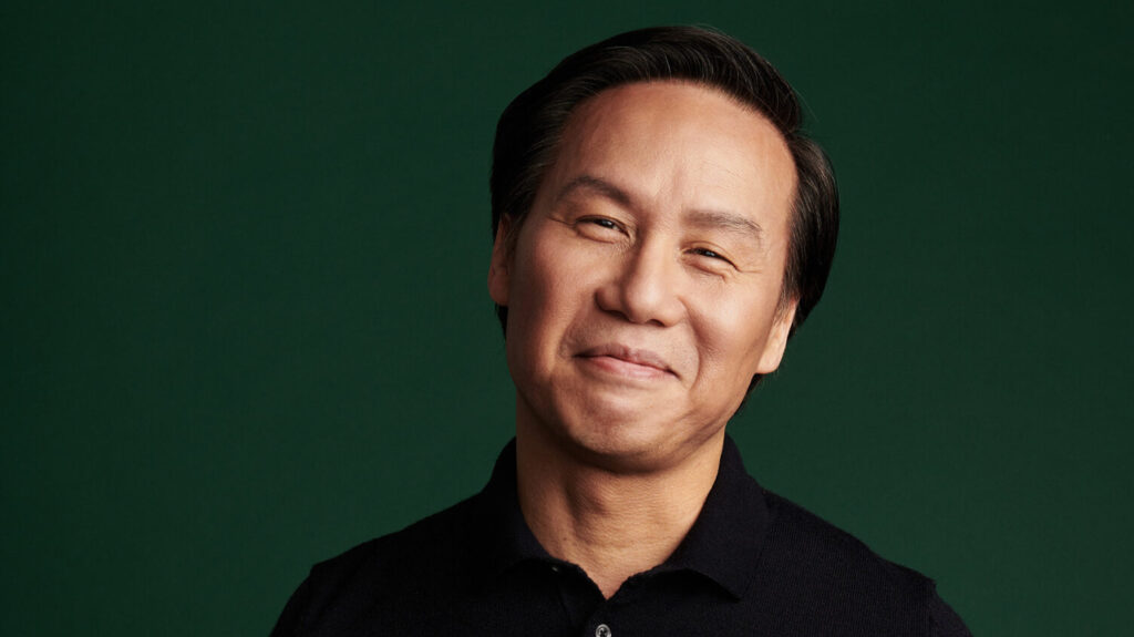 A confident man with a subtle smile, dressed in a black shirt against a dark green background.