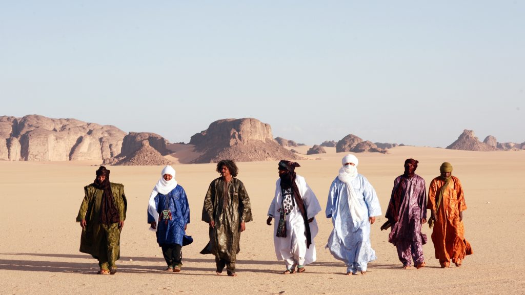 A diverse group of people in traditional attire walking together across a vast desert with striking rock formations in the background.