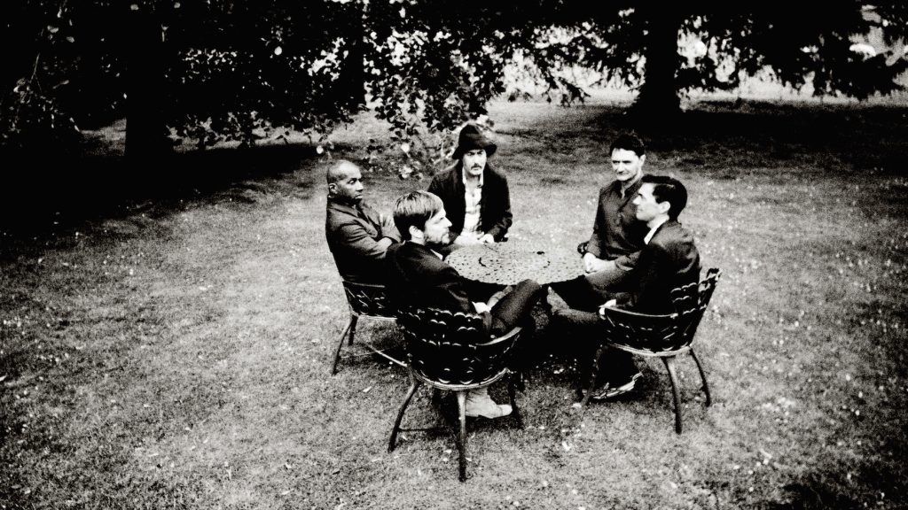 A group of friends engaged in an outdoor conversation, seated around a wrought iron table beneath the canopy of trees, in a moment that captures a relaxed and timeless camaraderie.