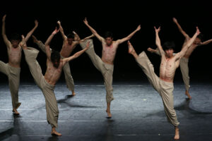 A group of male dancers performing a synchronized contemporary dance routine on stage, showcasing their strength and flexibility under dramatic lighting.