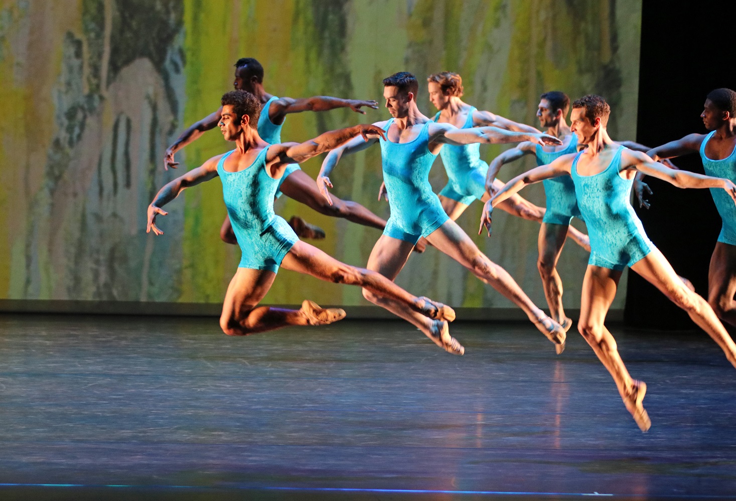 Seven male dancers in bright turquoise leotards leap across a stage in synchronized ballet positions against a muted yellow and green background.