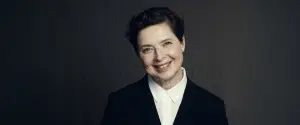 Smiling person with short hair wearing a black blazer and white shirt against a dark background.