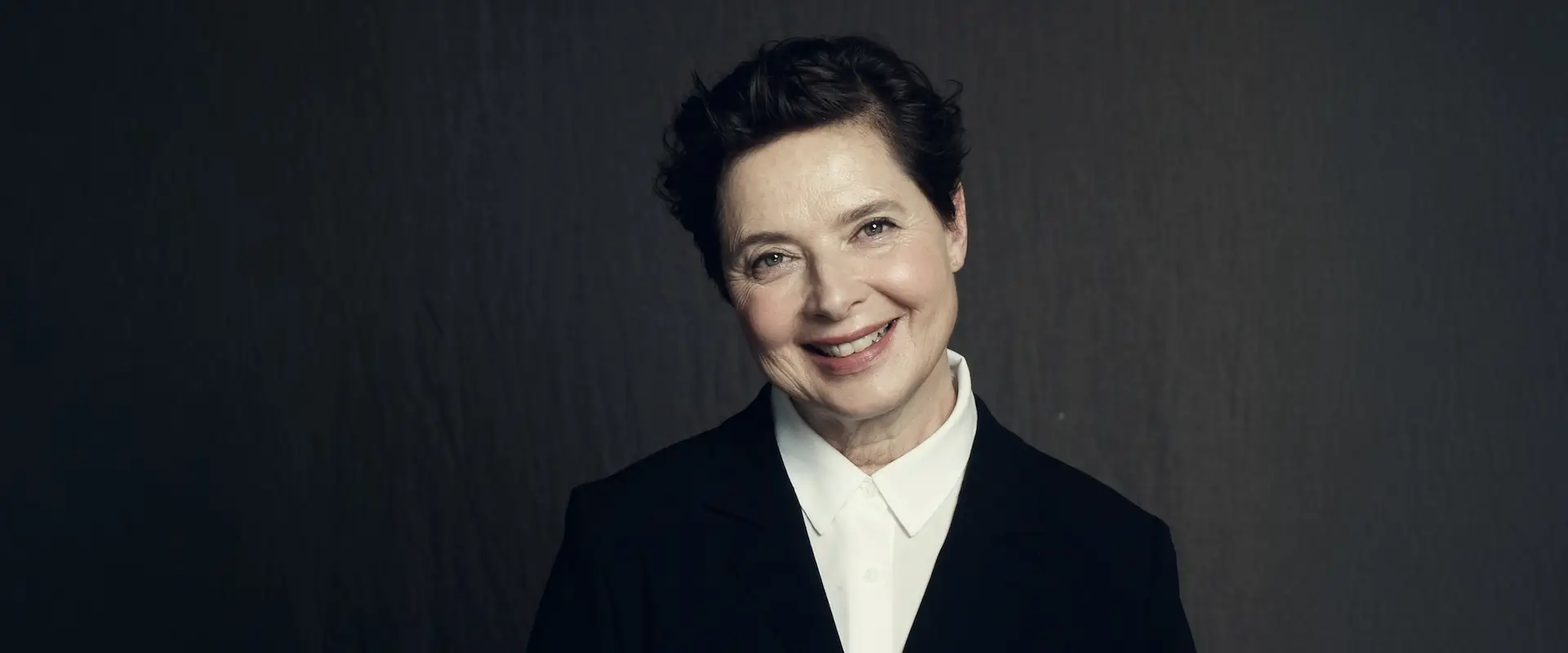 Smiling person with short hair wearing a black blazer and white shirt against a dark background.