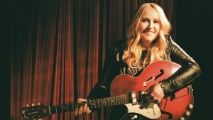 Musician with a radiant smile holding a red electric guitar, basking in the spotlight with a warm, curtain backdrop setting the stage for an intimate performance.