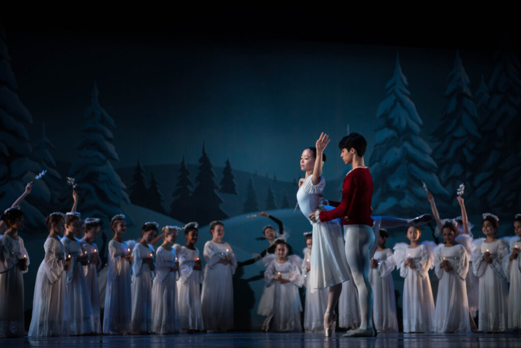 A ballet performance: a male and female lead dancer hold hands center stage, surrounded by a corps de ballet, against an enchanting winter backdrop.