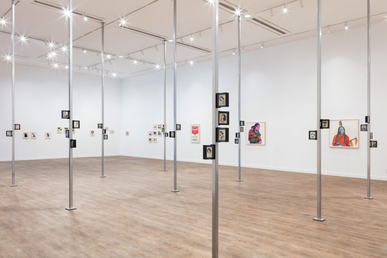 The Andy Warhol exhibition interior showcasing a diverse collection of framed artworks hung on white walls, with a polished wooden floor and an industrial-style ceiling fitted with track lighting.