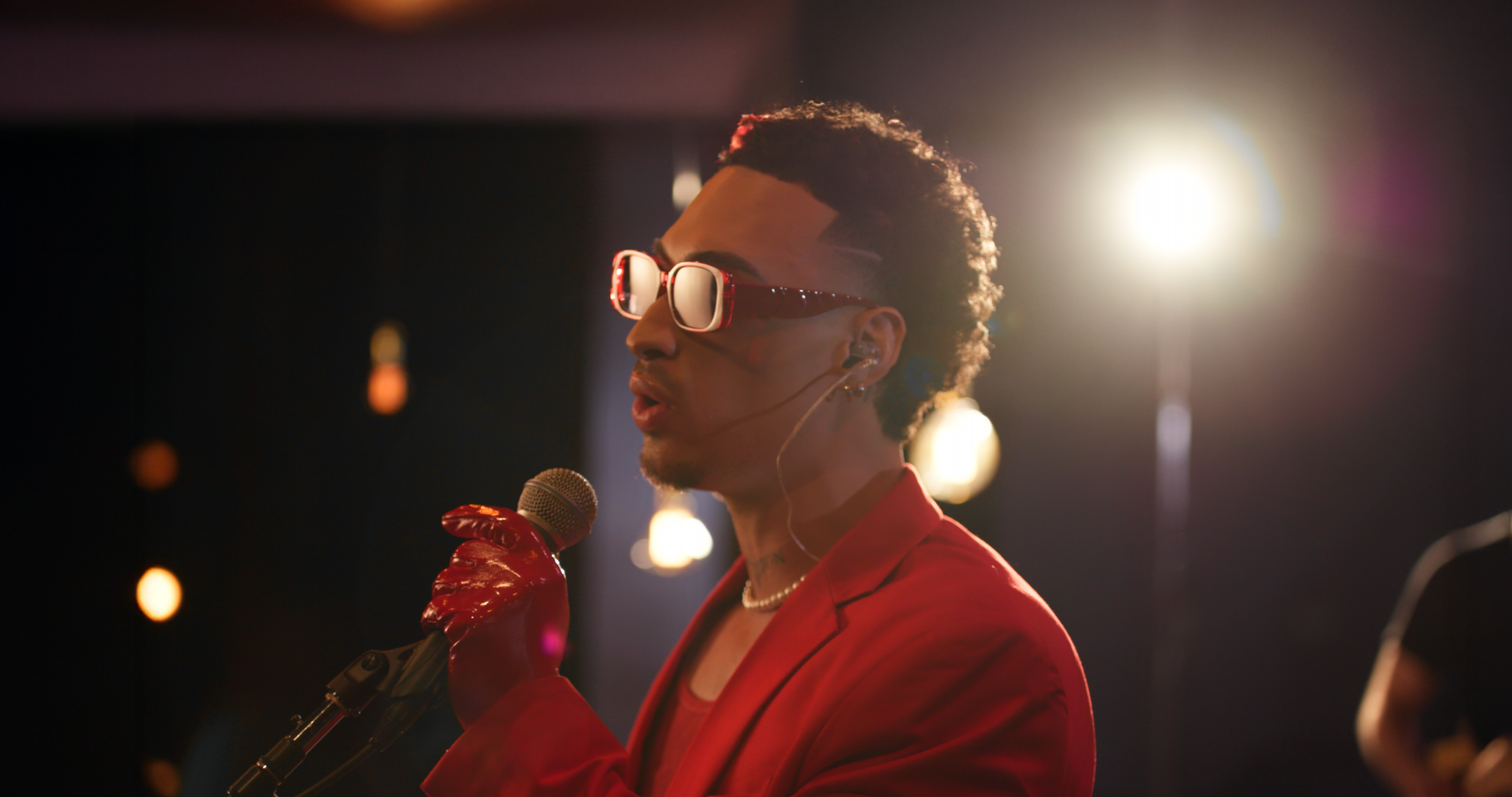 Lorenzo, in red with sunglasses and gloves, sings into a microphone under dramatic stage lights.