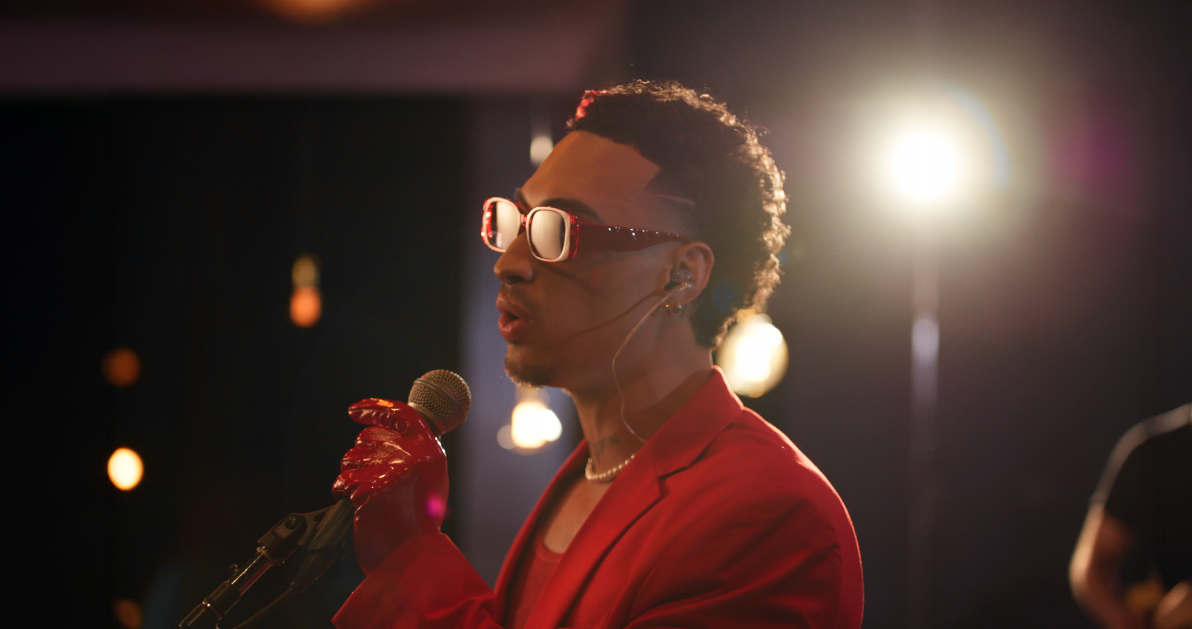 A person wearing sunglasses, red gloves, and a red outfit is singing into a microphone. They have a focused expression. Bright stage lights are visible in the background, creating a dramatic atmosphere.