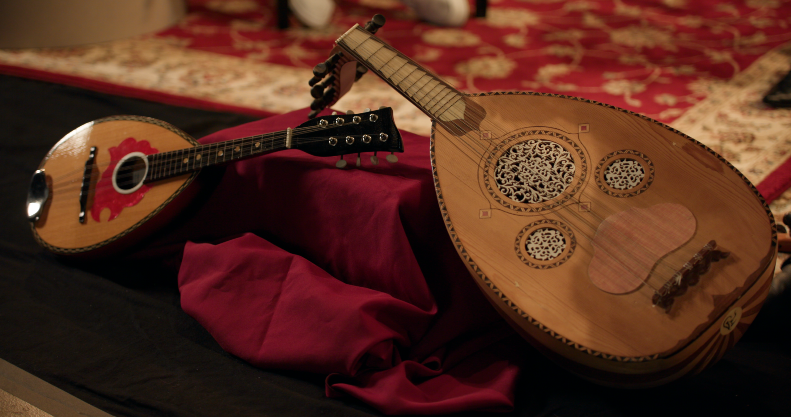A mandolin and oud rest on red cloth, with a patterned red carpet in the background.