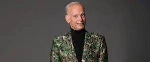 A person with short gray hair wears a green patterned jacket over a black turtleneck, standing against a gray background.