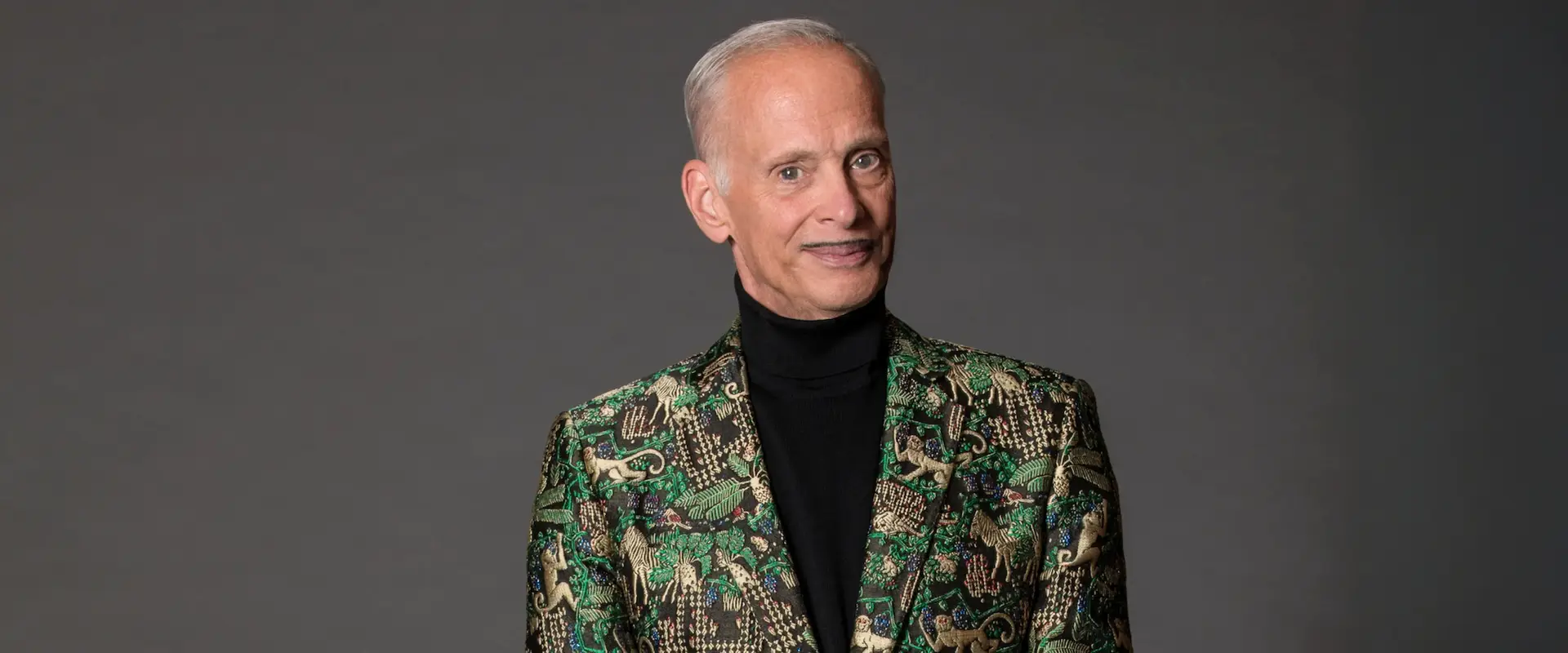 A person with short gray hair wears a green patterned jacket over a black turtleneck, standing against a gray background.