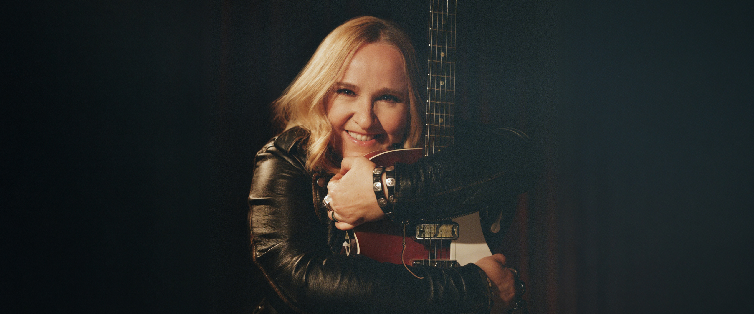 Melissa Etheridge with blonde hair smiles, hugging an electric guitar in a black leather jacket.