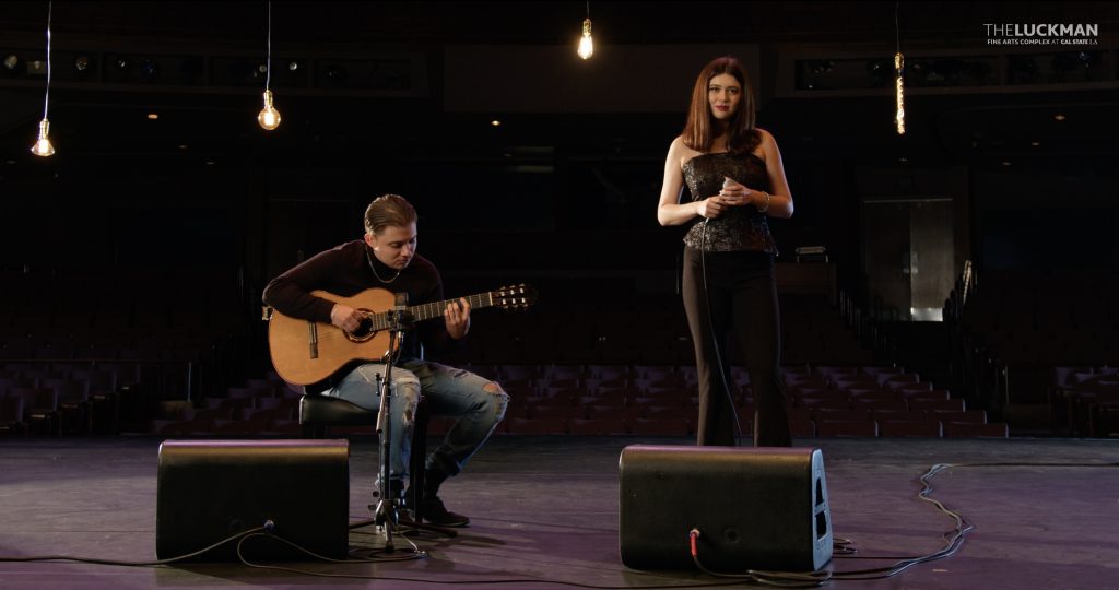Two musicians practicing on stage, with a guitarist seated and playing an acoustic guitar while a vocalist stands beside with a microphone, set against the backdrop of an empty concert hall.