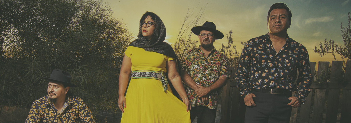 La Santa Cecilia group posing with confidence in a rustic outdoor setting at dusk, exuding a vintage vibe.
