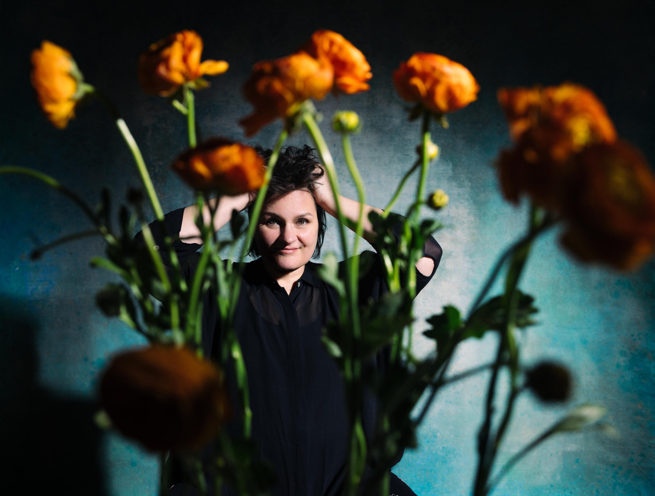 Madeline Peyroux smiling through an array of vibrant orange flowers with a dark blue background.