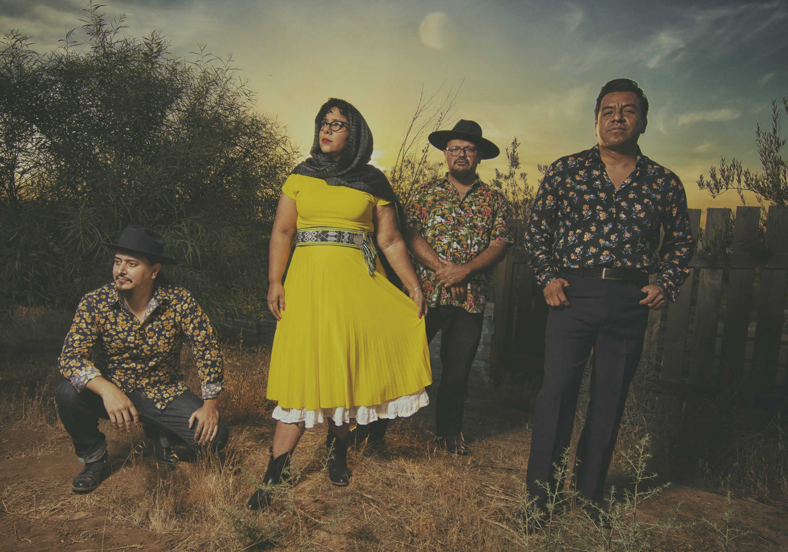 La Santa Cecilia group posing with confidence in a rustic outdoor setting at dusk, exuding a vintage vibe.