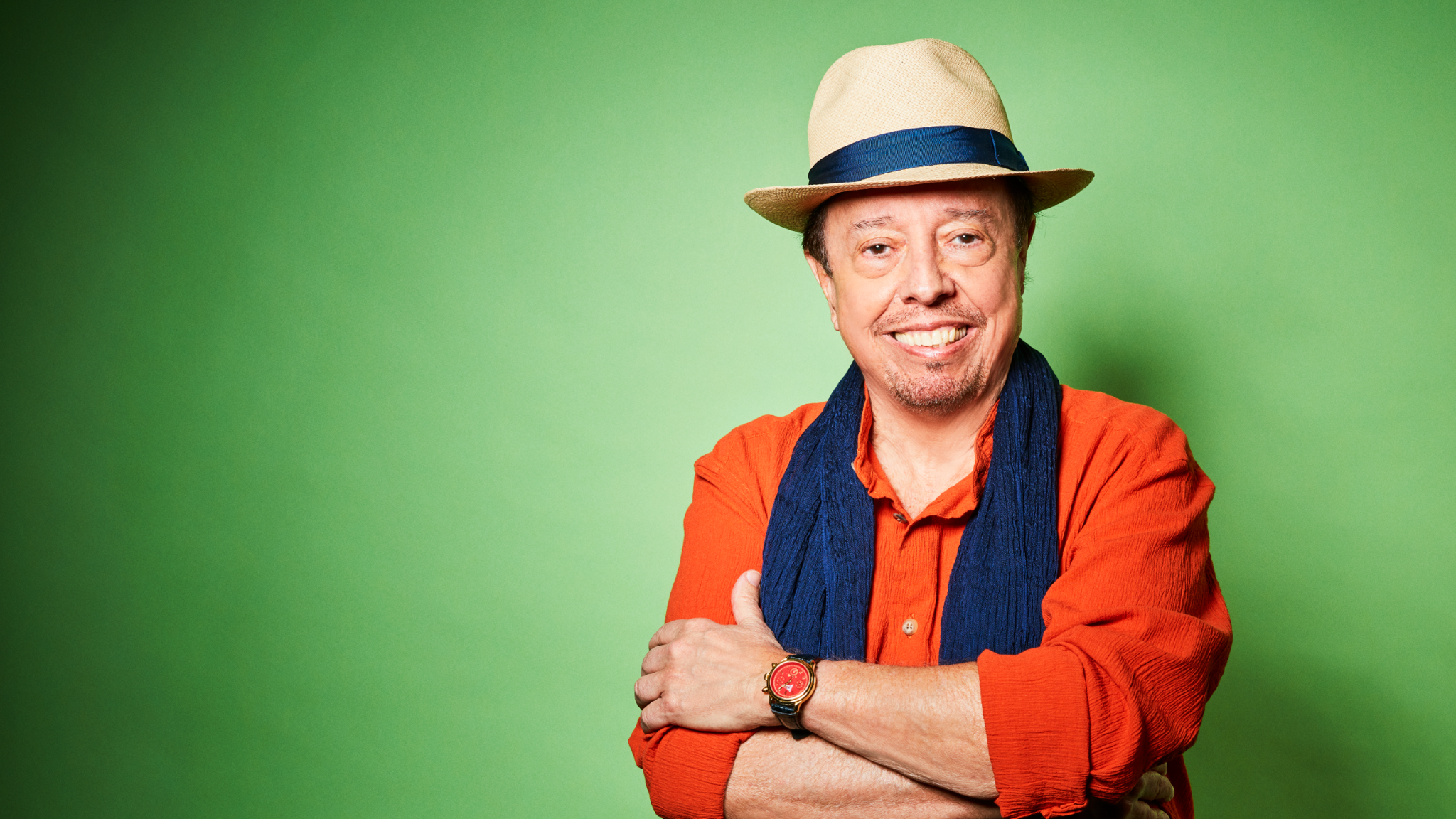 Sergio Mendes with a beaming smile posing confidently against a green background, dressed in a vibrant orange shirt, blue scarf, and a stylish straw hat, exuding charisma and joy.