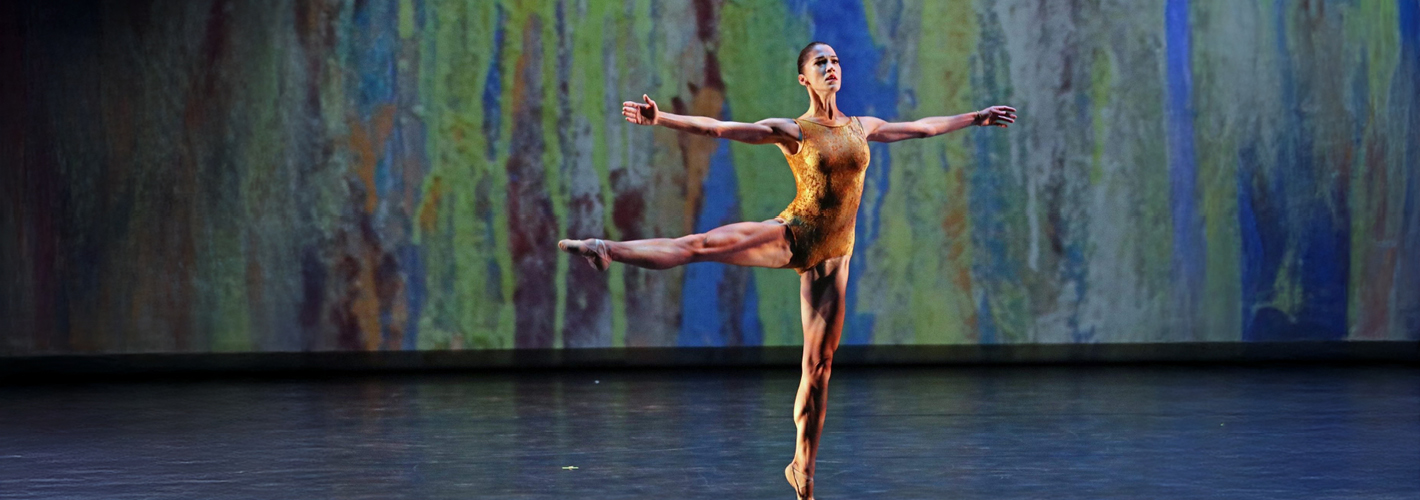 A ballet dancer, Tetyana Martyanova, in a gold costume balances en pointe with one leg extended and arms outstretched against a colorful, abstract stage backdrop.