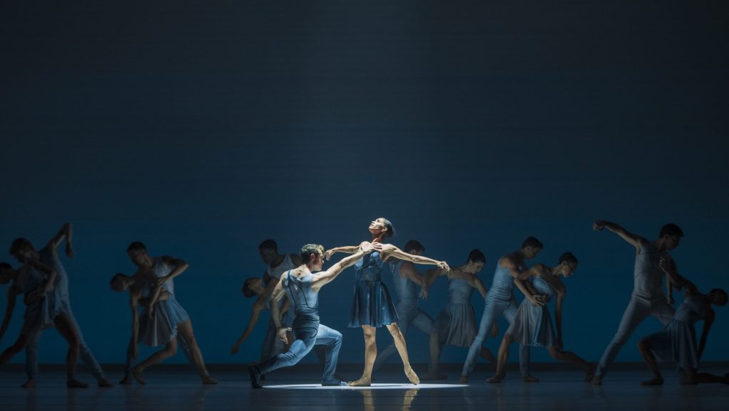 Elegant ballet performance of La Compañía Nacional de Danza with a male and female dancer in a duet spotlight, surrounded by a corps of ballet in synchronized grace against a moody blue backdrop.