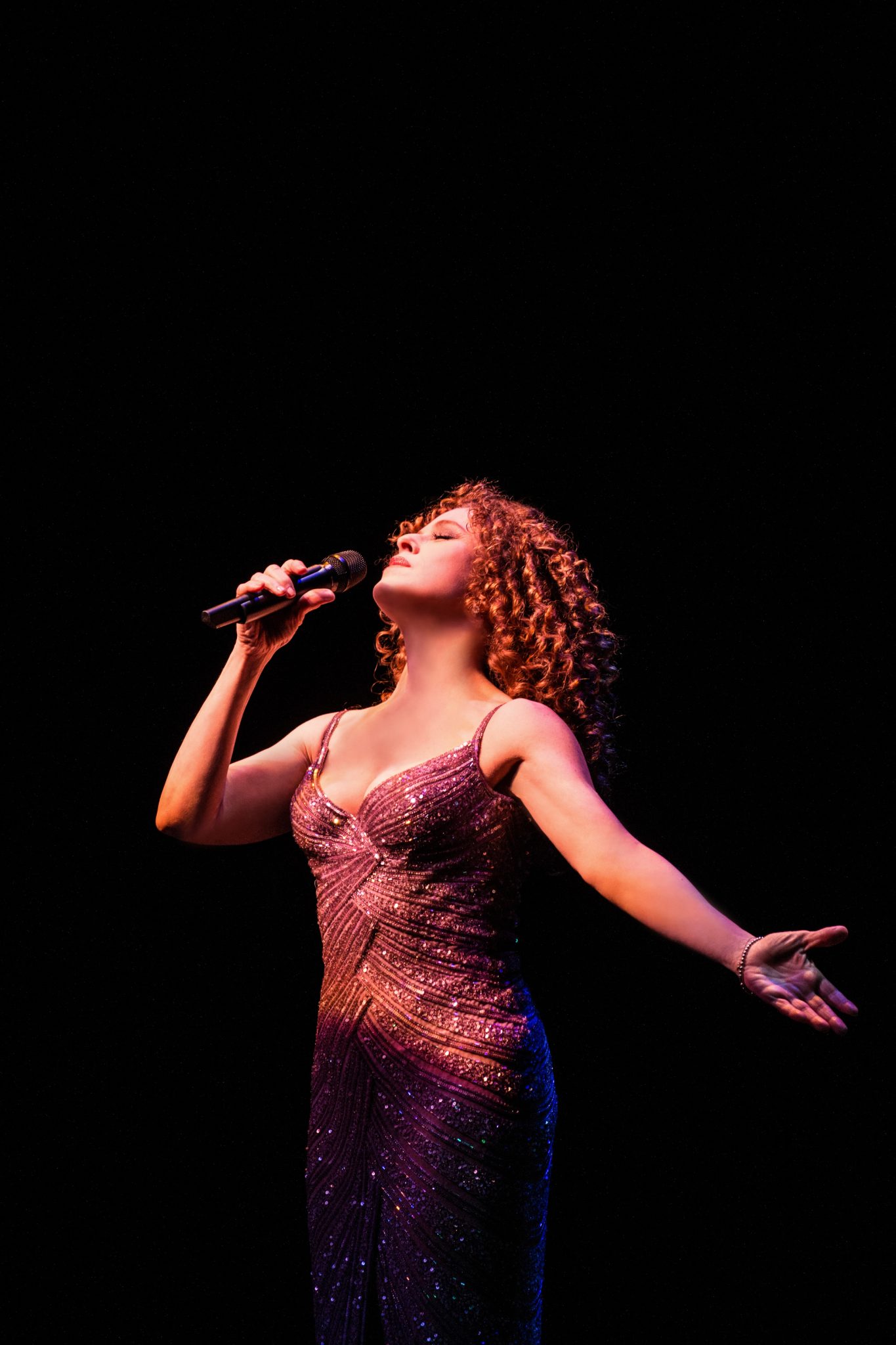 Bernadette Peters singing passionately into a microphone on a dark stage, spotlight highlighting her expressive stance.