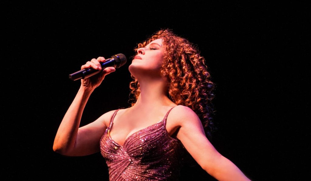 Bernadette Peters singing passionately into a microphone on a dark stage, spotlight highlighting her expressive stance.