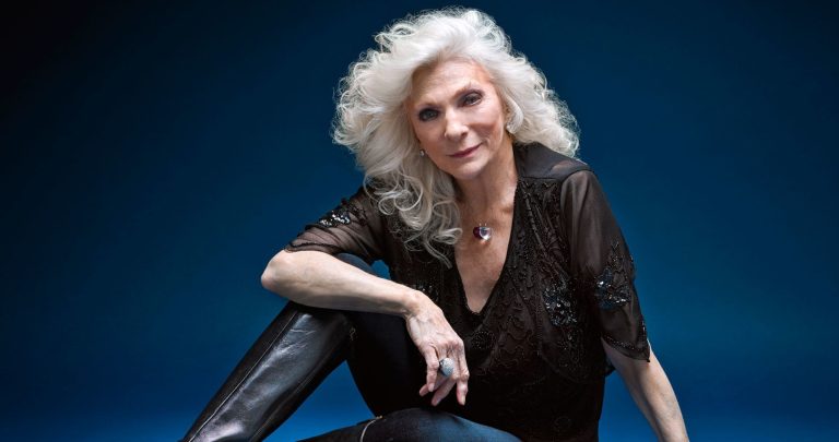 Elegant Judy Collins with silver hair posing confidently against a blue backdrop.