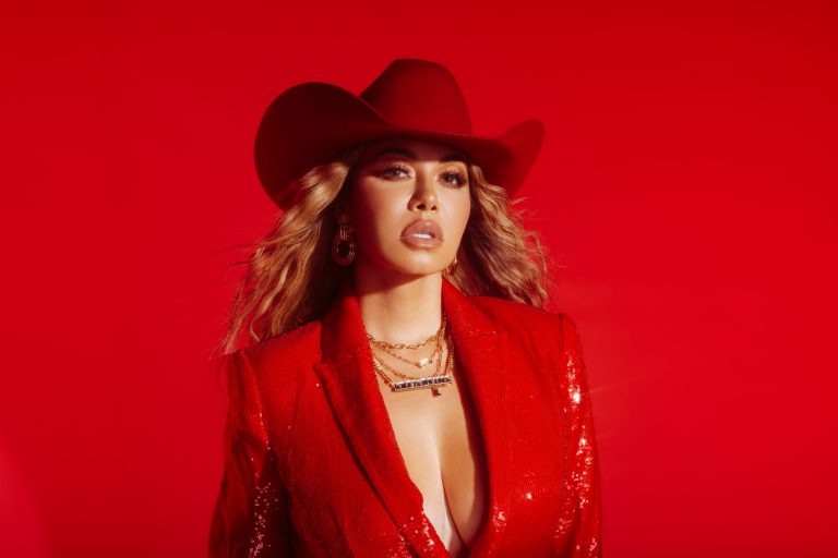 Chiquis in a stylish red sequined suit and matching wide-brimmed hat poses confidently against a vibrant red background, exuding glamour and fashion-forward flair.