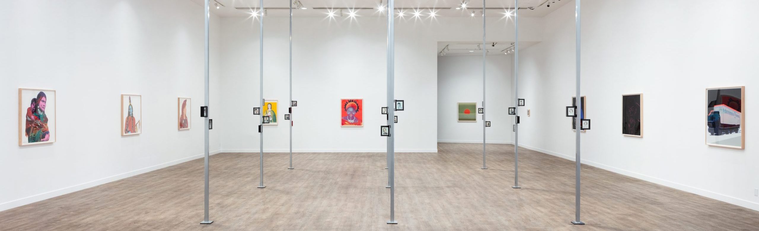 A spacious art gallery interior hosting an Andy Warhol exhibition, featuring a varied collection of framed artworks on white walls and a series of standing informational placards, under a modern lighting system.