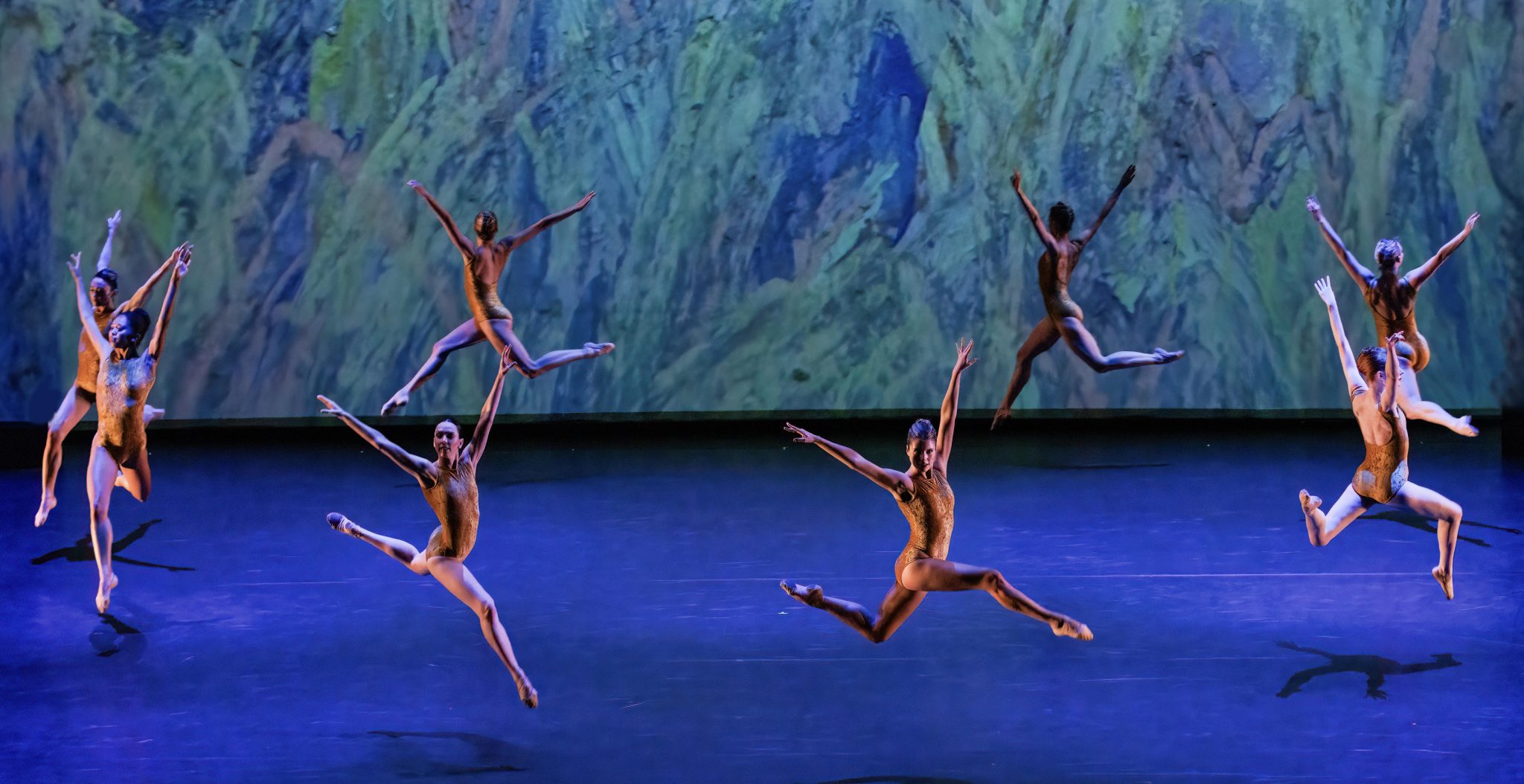Raiford Rogers ballet dancers in perfect unison mid-jump against a painted backdrop, exhibiting the artistry and athleticism of a dance performance.