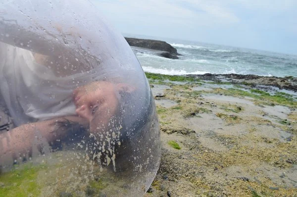 A person peering through a translucent, water-splattered orb on a rocky beach with waves crashing in the background.