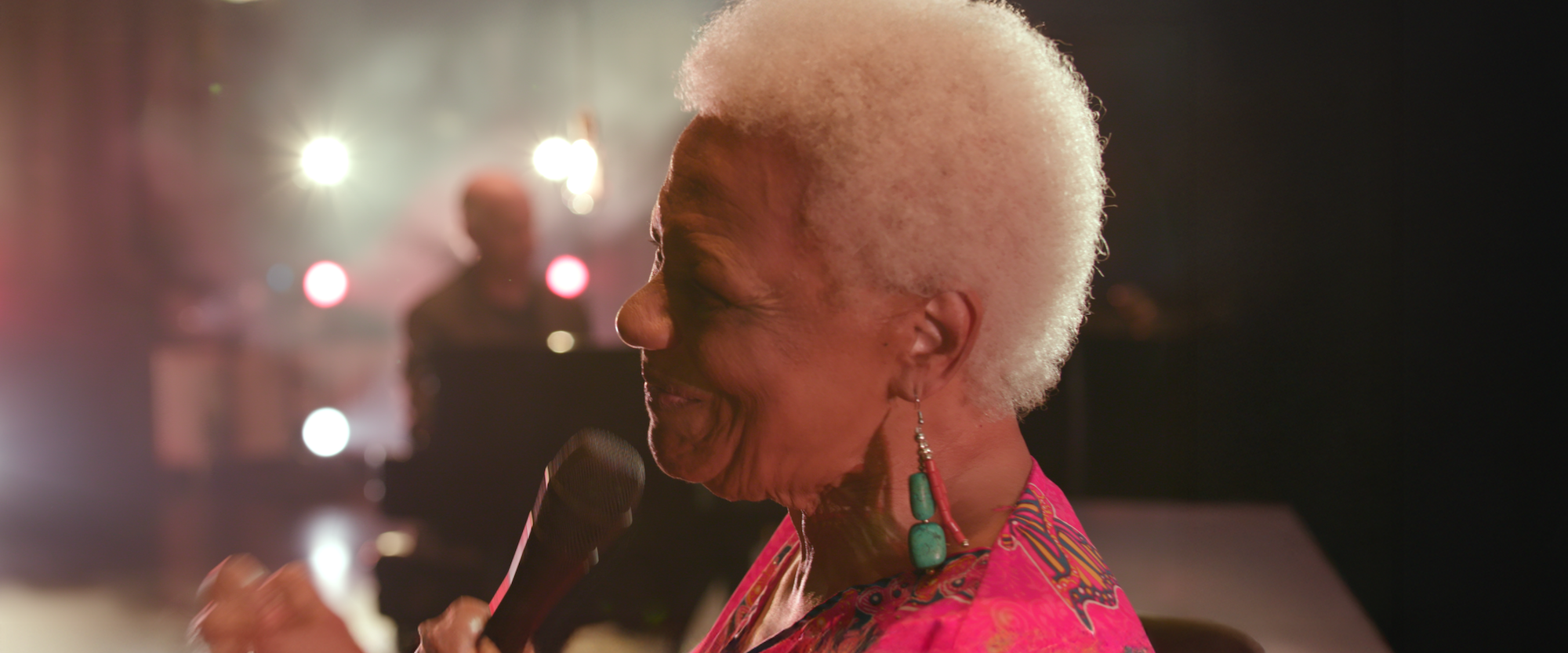 A woman with short white hair, wearing bright clothing and turquoise earrings, holds a microphone and smiles, with colorful stage lights blurred in the background.