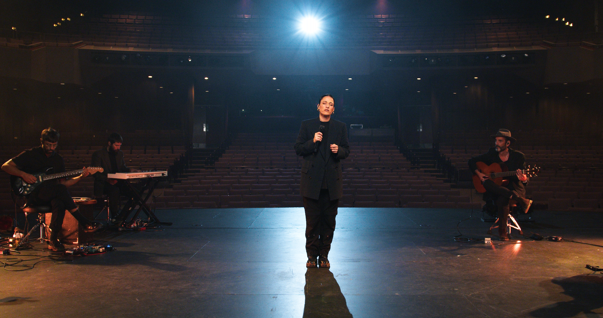 A performer stands on a dimly lit stage with a spotlight shining from above. They hold a microphone and wear a black outfit. Three musicians sit nearby playing guitar and keyboards. The backdrop reveals an empty auditorium.