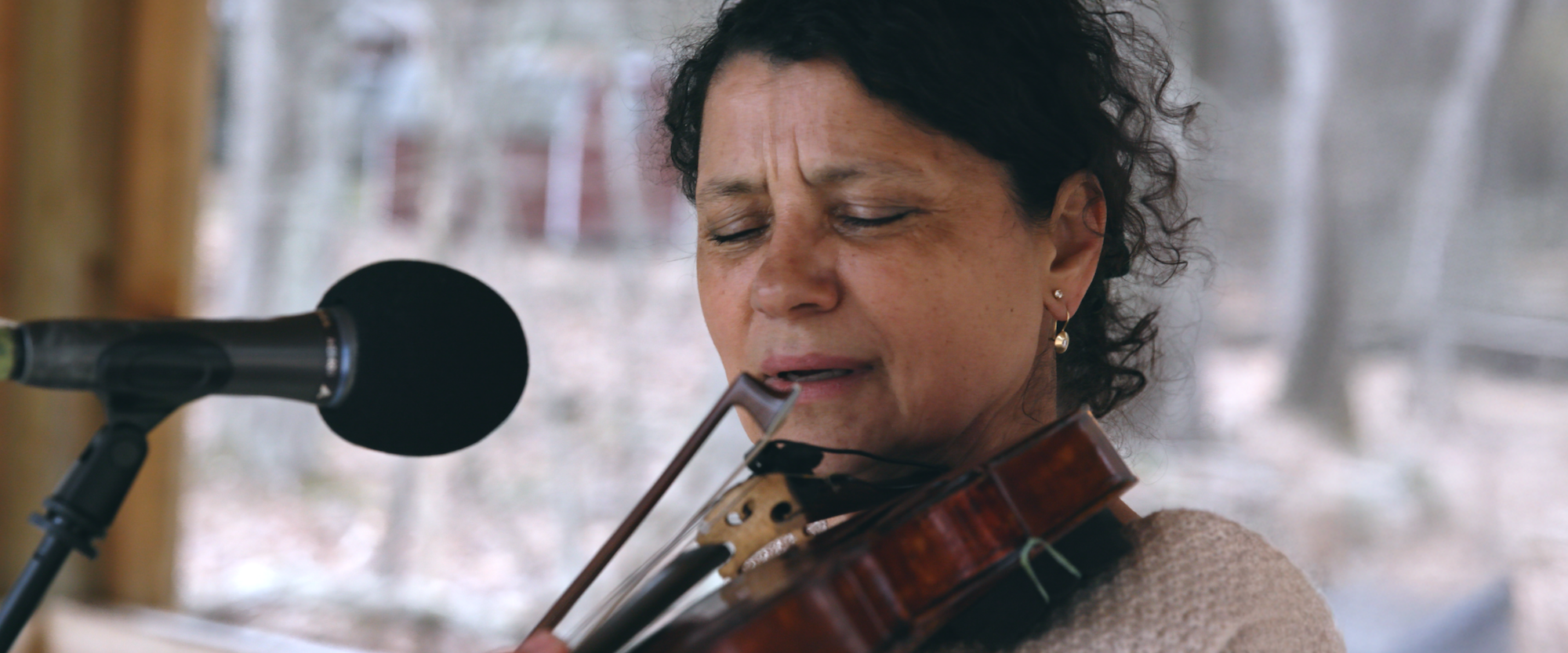 Iva Bittova playing a violin with eyes closed, wearing a beige sweater and gold earrings. She is in front of a microphone, and the background is softly blurred. Her expression is focused and serene.